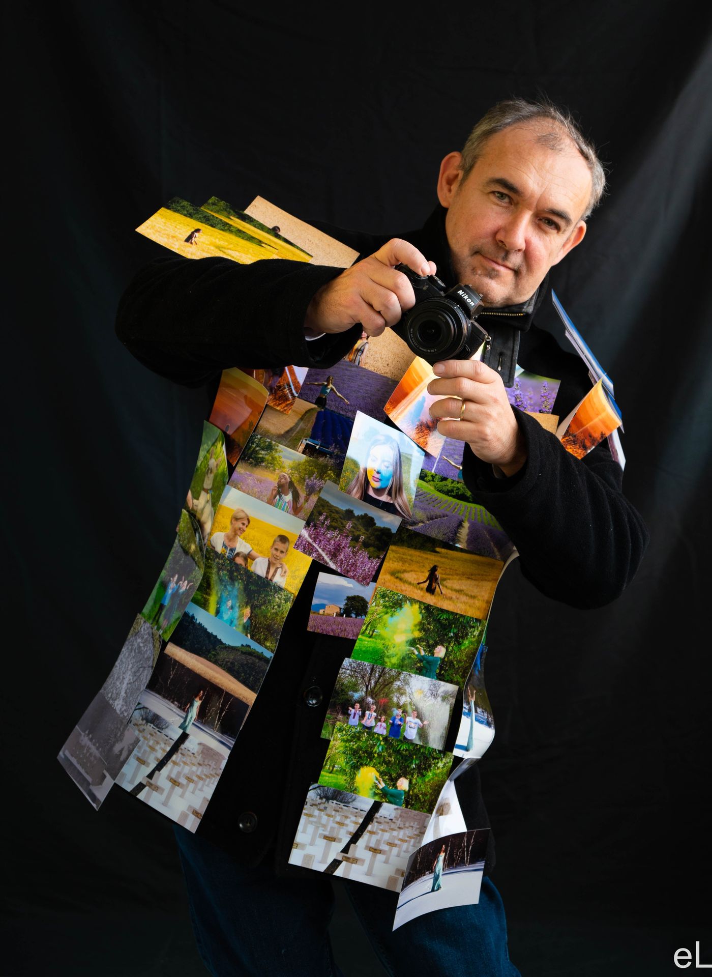 Le photographe, appareil levé, vêtu d'une veste recouverte de tirages de ses propres photographies.