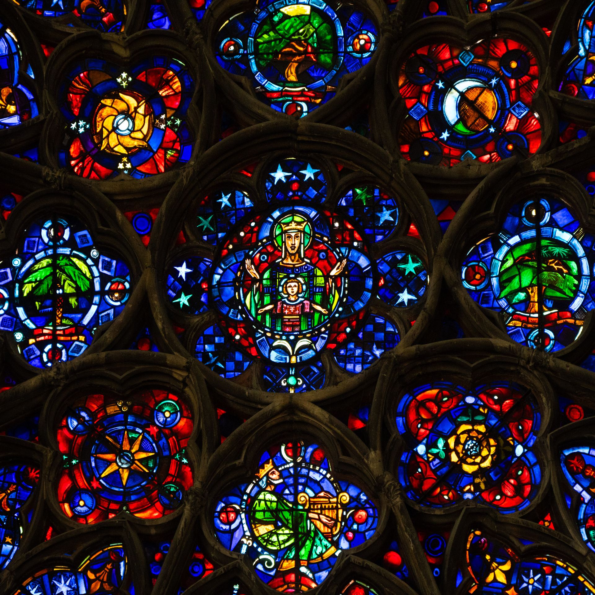 Une rosace de cathédrale en vitrail, la Vierge à l'Enfant en son centre, parmi les étoiles, les palmes et les bleus et rouges profonds.